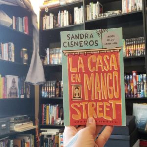 A hand holding the book La Casa En Mango Street in front of bookshelves